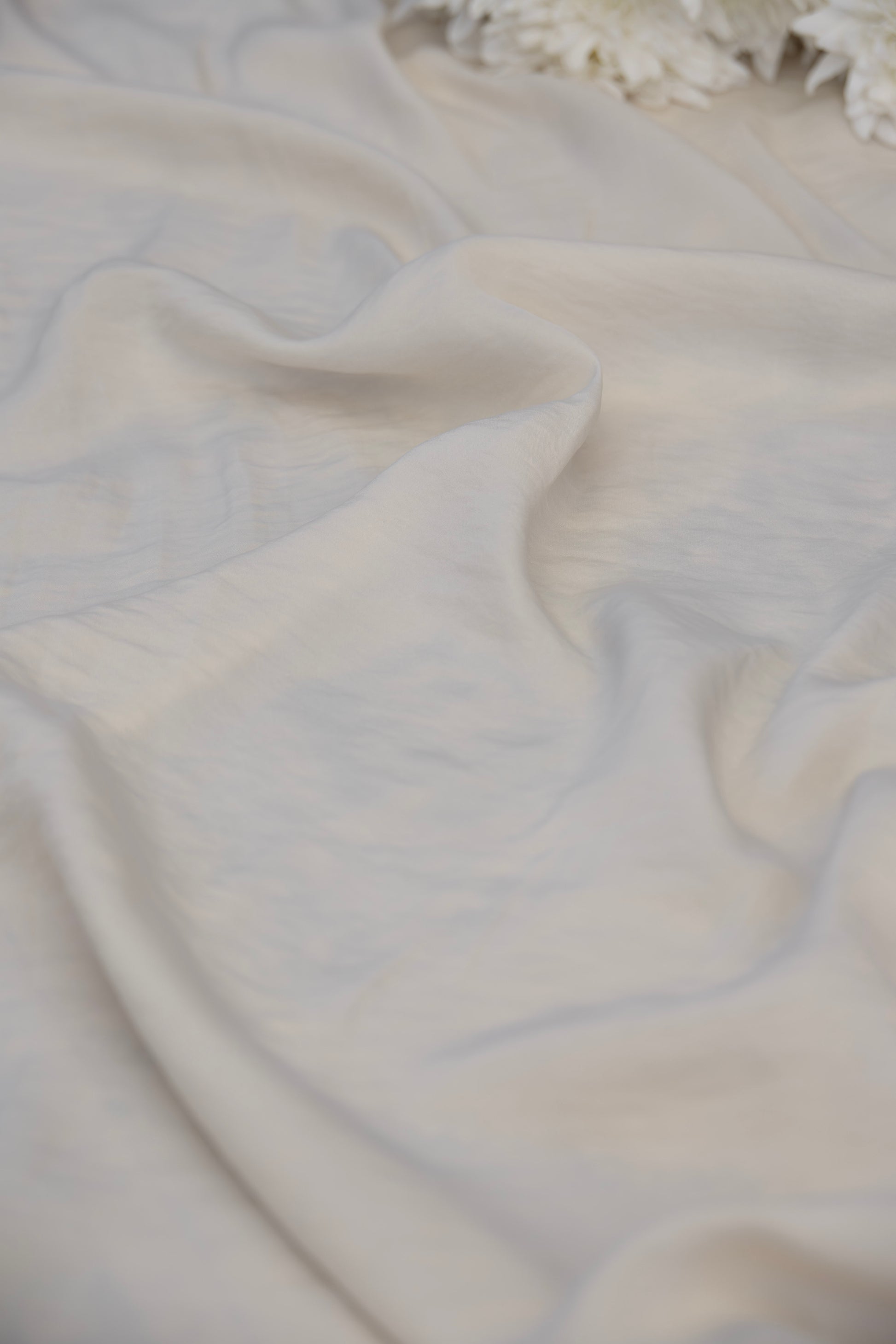 A close-up of draped off-white fabric with soft folds and a slightly textured surface, with a small portion of white flowers visible at the top edge.