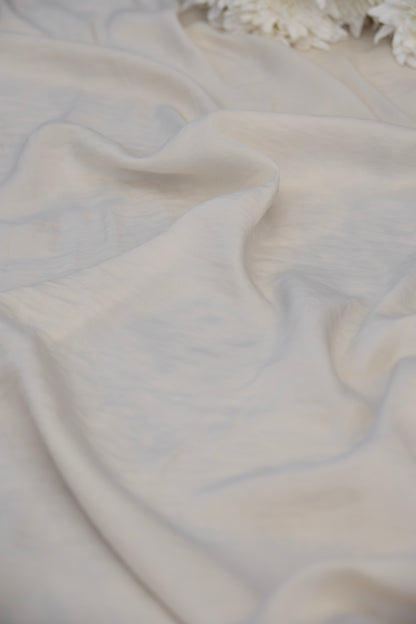 A close-up of draped off-white fabric with soft folds and a slightly textured surface, with a small portion of white flowers visible at the top edge.
