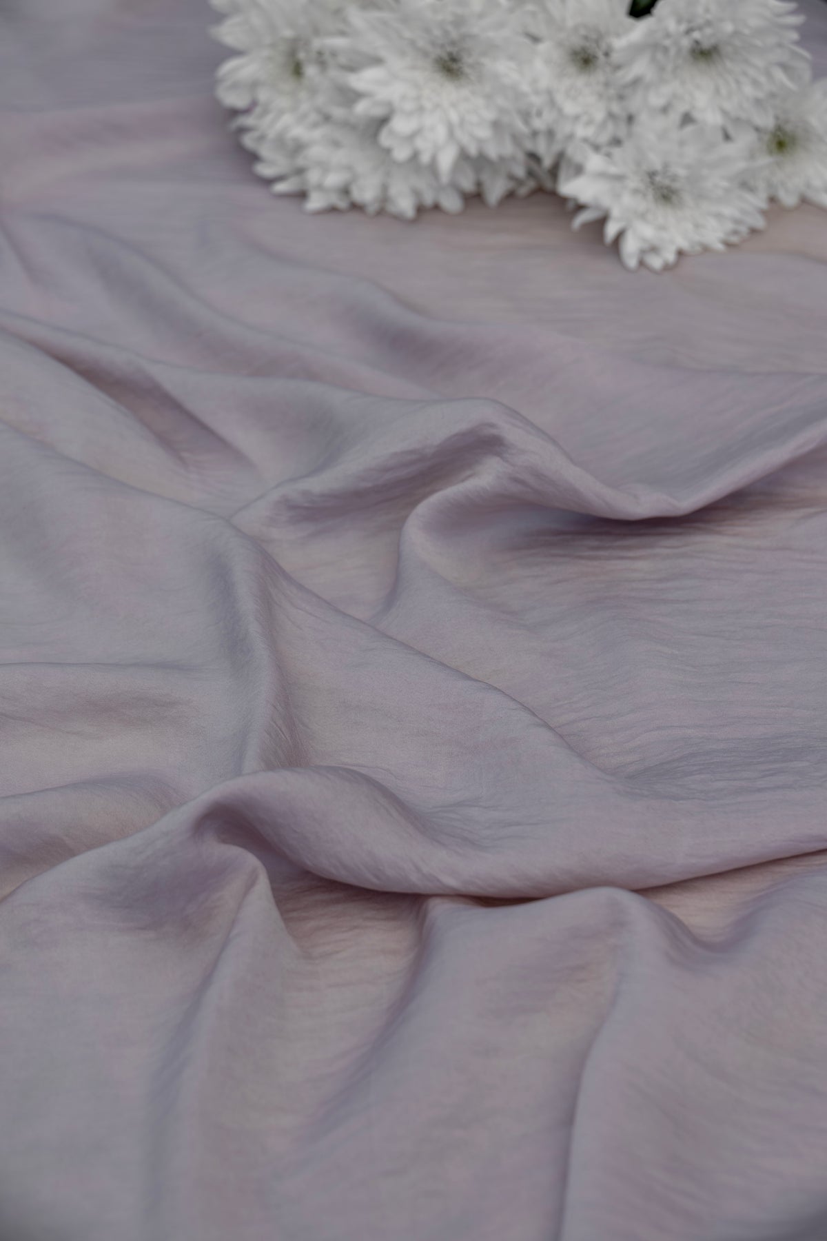 A close-up of draped dusty lavender fabric with soft folds, with a small cluster of white flowers placed in the upper right corner.