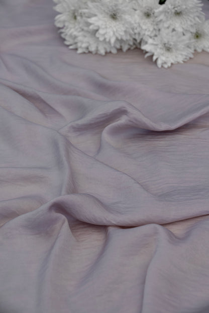 A close-up of draped dusty lavender fabric with soft folds, with a small cluster of white flowers placed in the upper right corner.