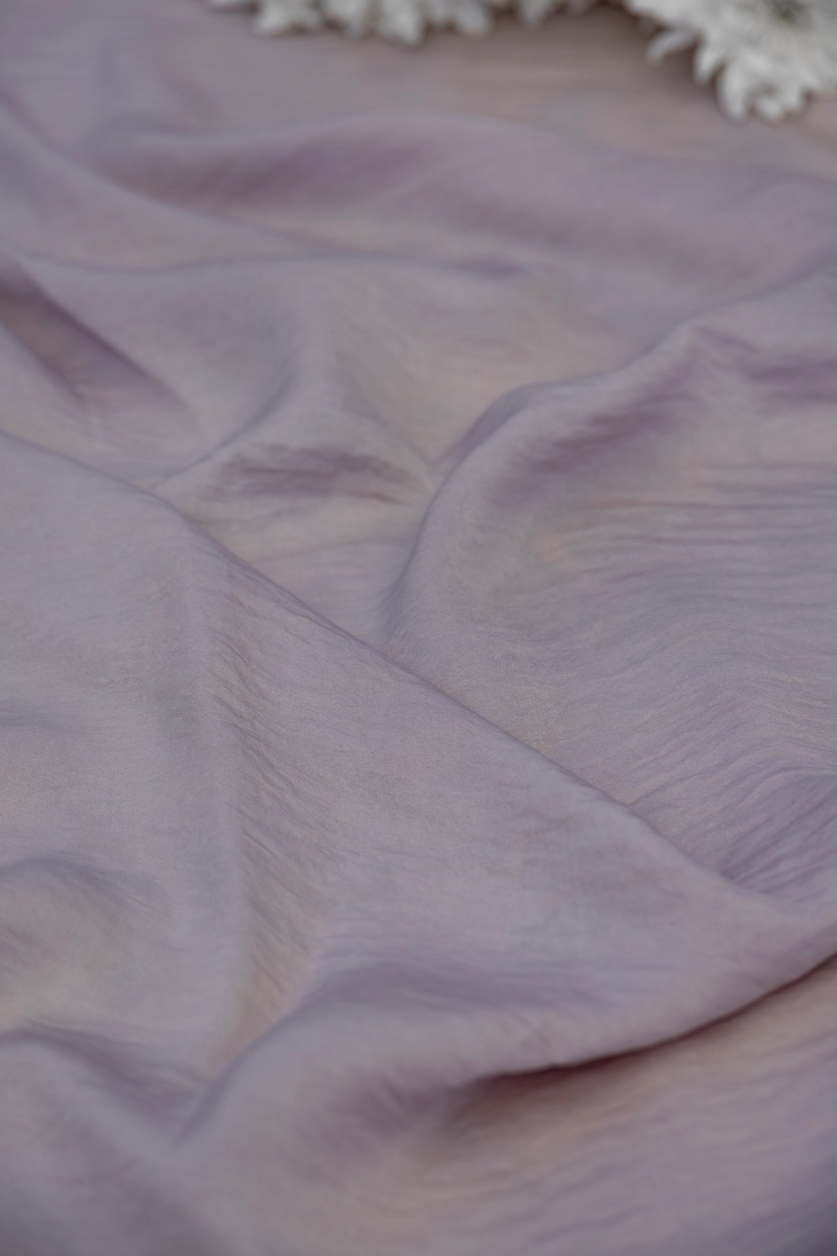 A close-up of draped lavender fabric with soft folds and a small portion of white floral decoration visible at the top edge.