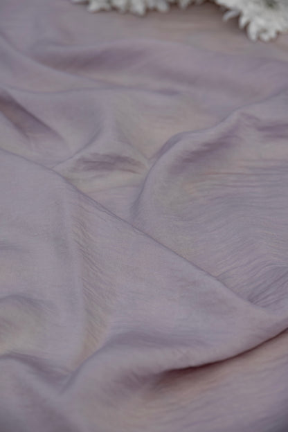 A close-up of draped lavender fabric with soft folds and a small portion of white floral decoration visible at the top edge.