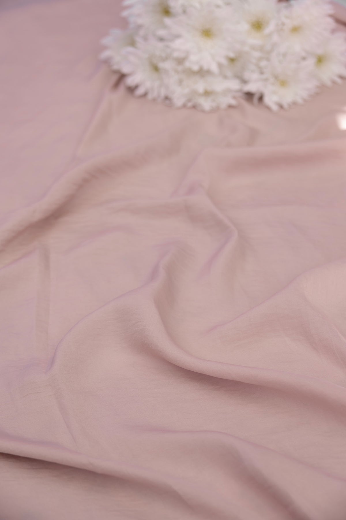 A close-up of draped dusty pink fabric with soft folds, topped near the top edge by a small cluster of white daisy-like flowers.