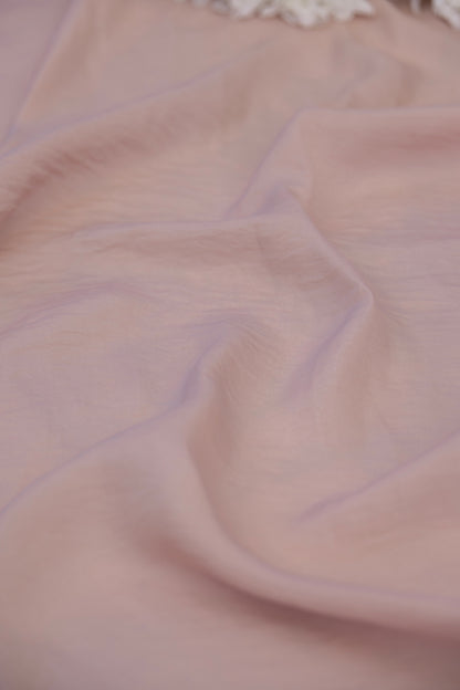 A close-up of draped light pink fabric with soft folds and a small portion of white lace or floral trim visible at the top edge.