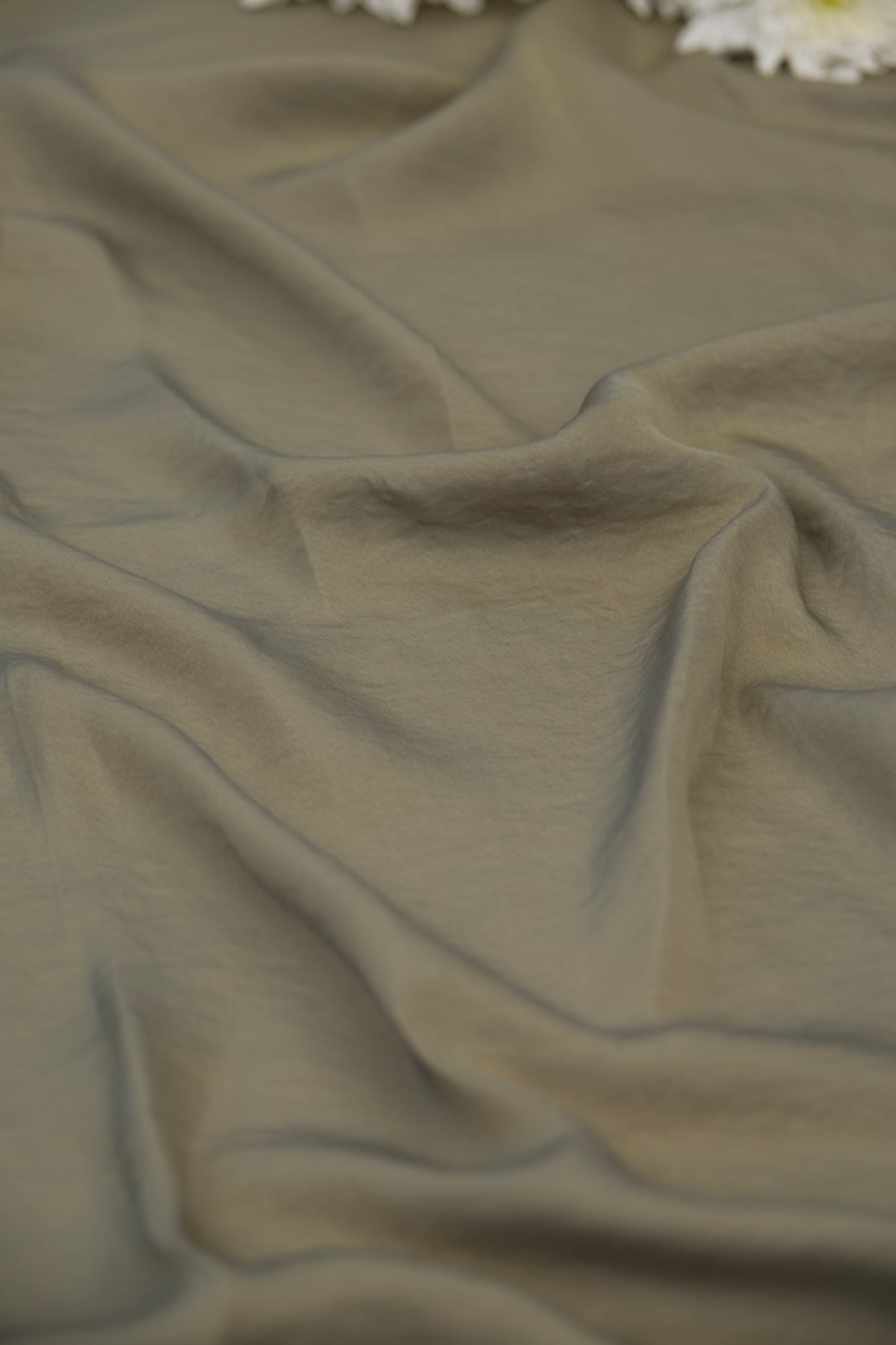 A close-up of draped olive green fabric with soft folds and a few white flowers visible at the top edge.