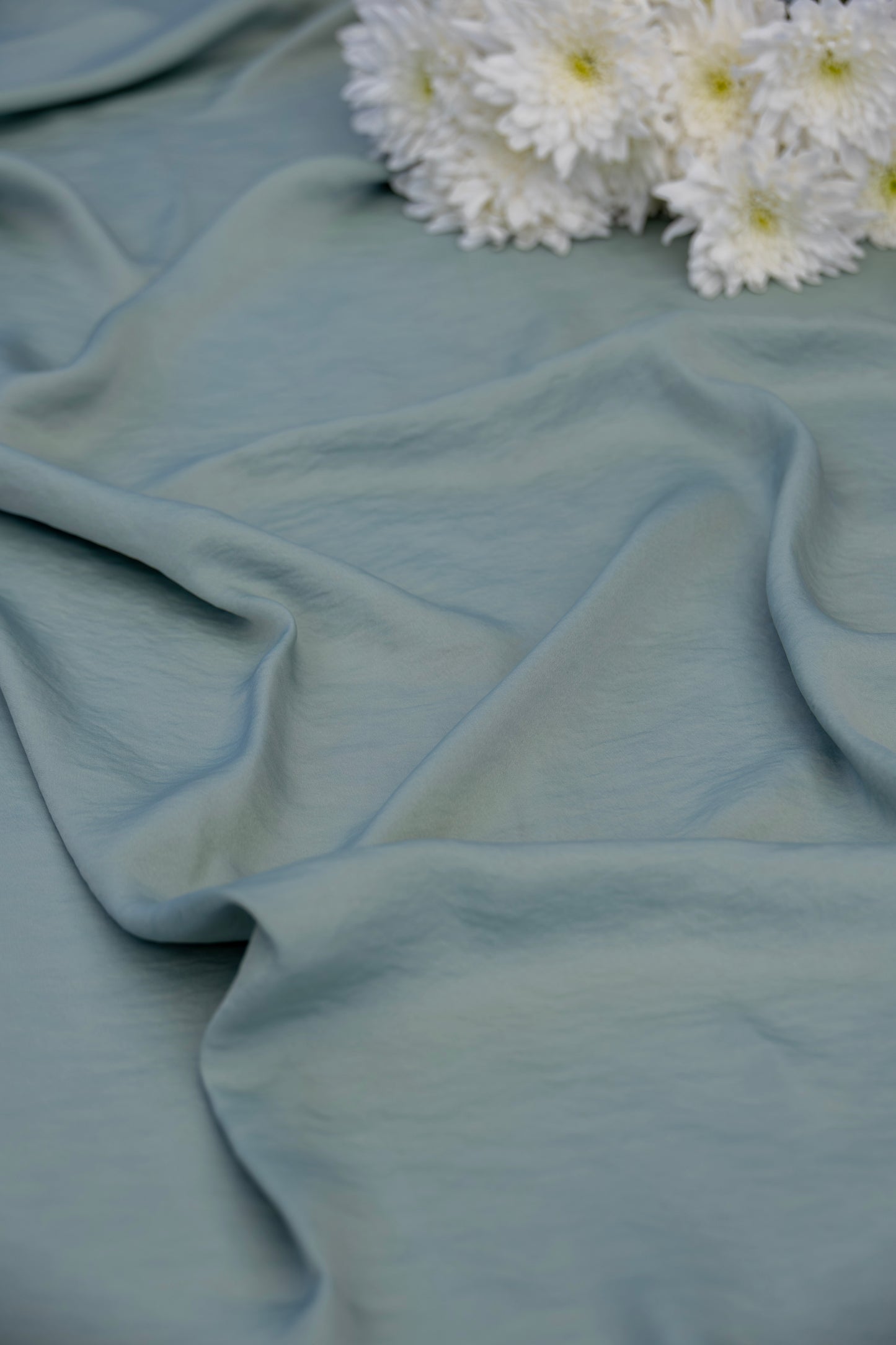 A close-up of draped dusty blue fabric with soft folds, styled with a small cluster of white daisy-like flowers in the upper right corner.