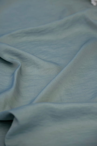 A close-up of draped dusty blue fabric with soft folds and a smooth texture.
