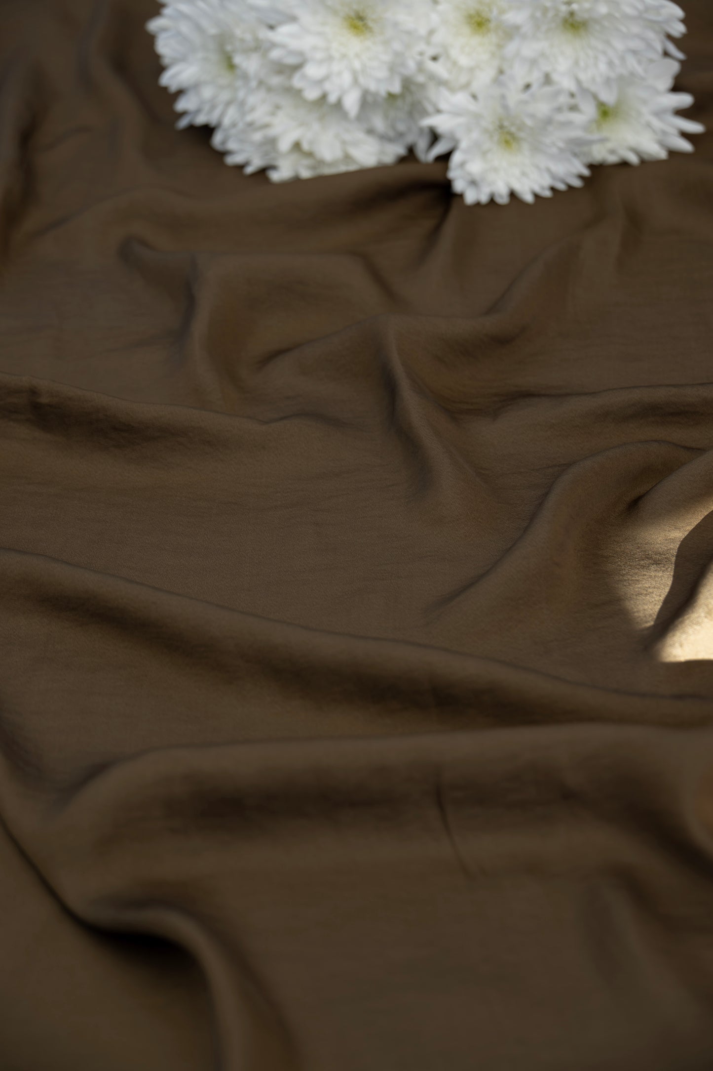 A close-up of draped brown fabric with soft folds and a small cluster of white flowers placed in the upper corner.