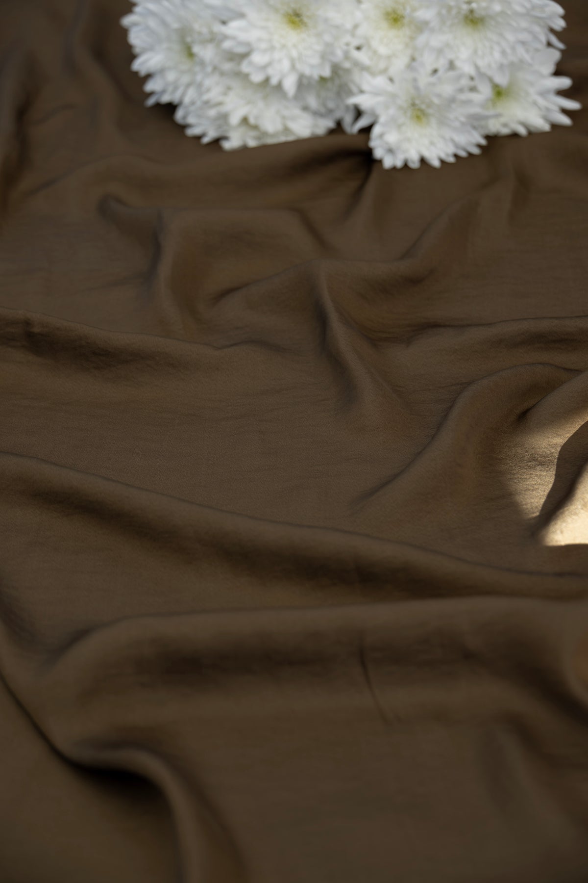 A close-up of draped brown fabric with soft folds and a small cluster of white flowers placed in the upper corner.