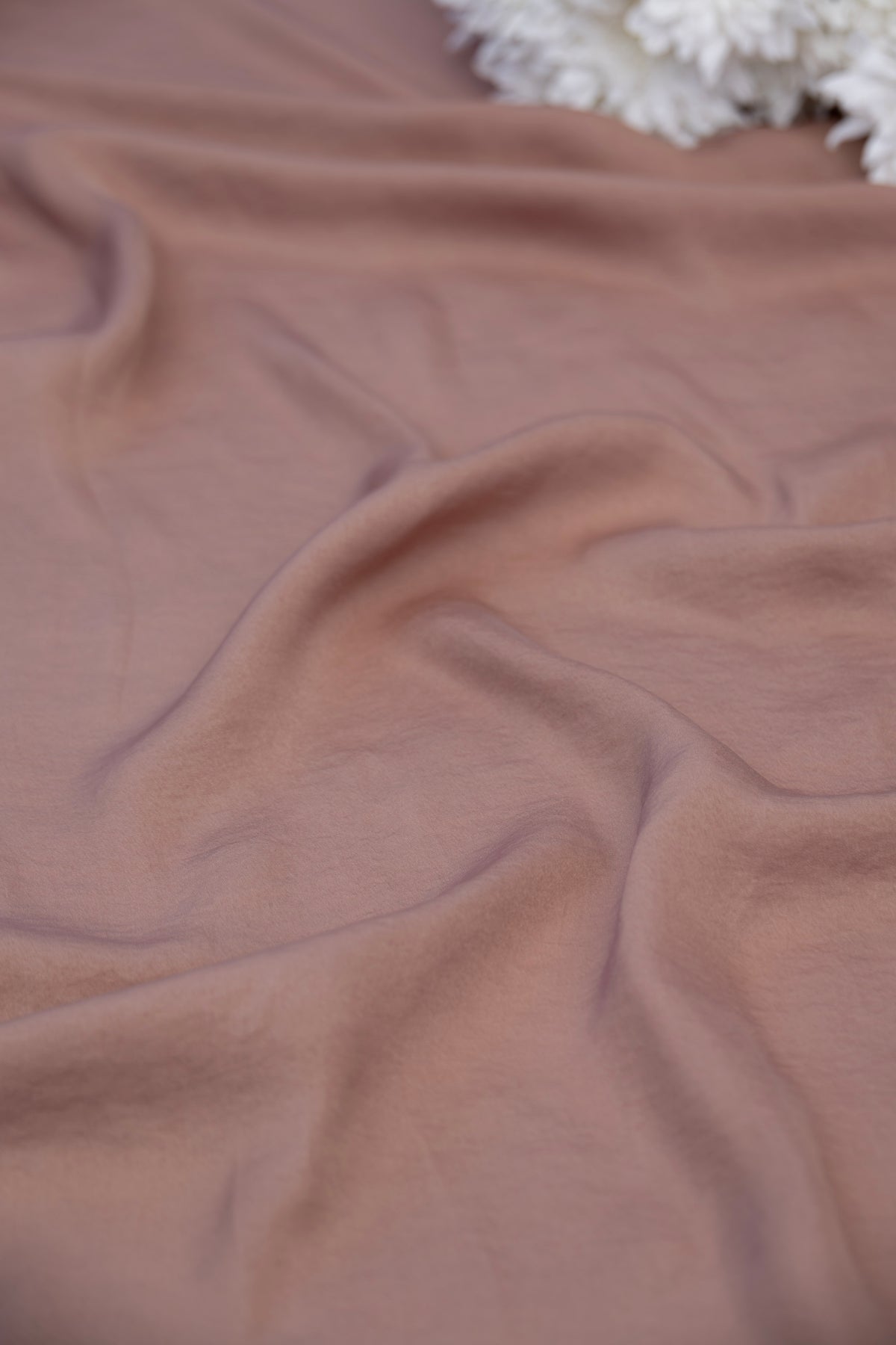 A close-up of draped dusty rose fabric with soft folds and a small portion of a white textured floral prop visible in the top right corner.