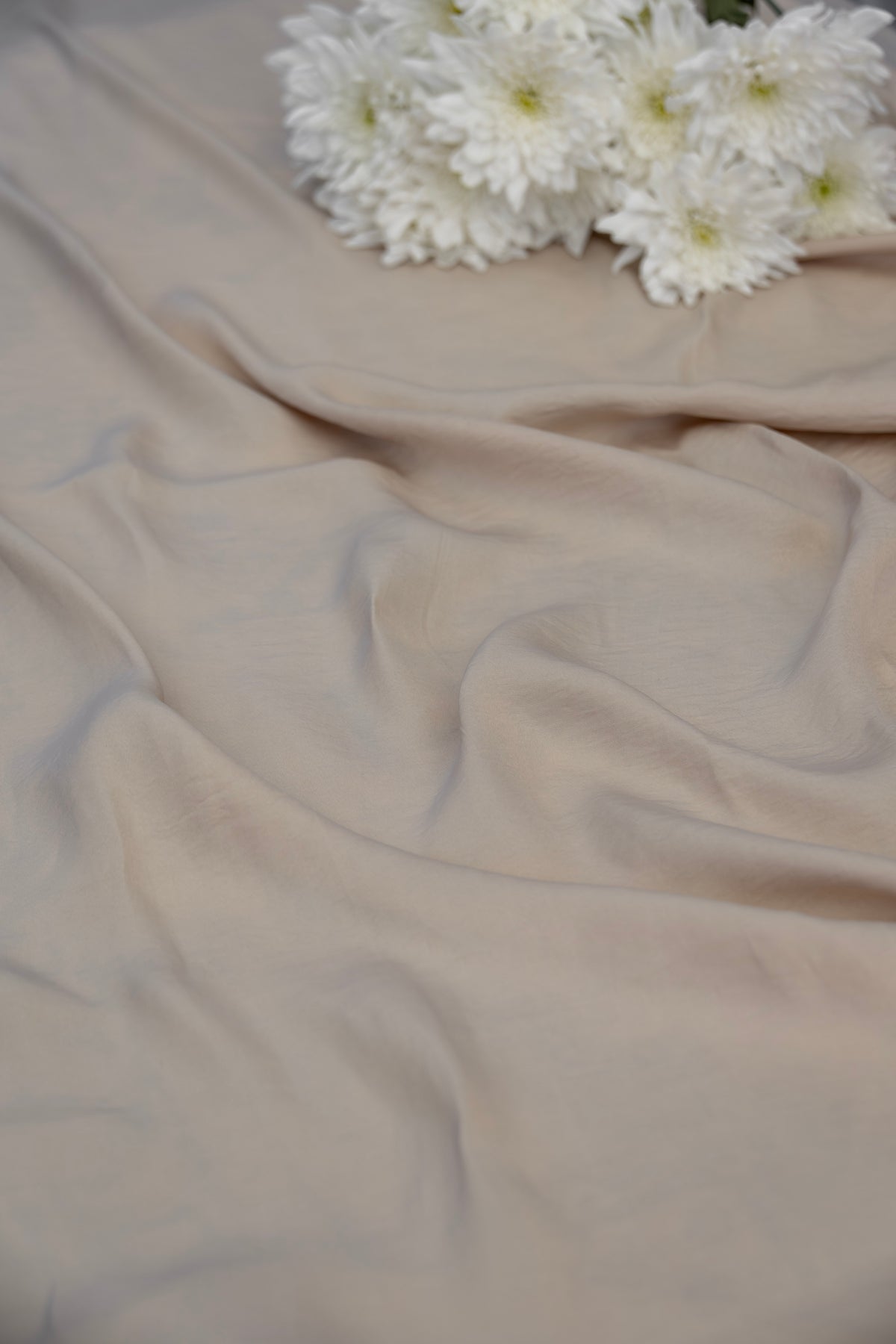 A draped piece of light beige fabric with soft folds, styled with a cluster of white chrysanthemum flowers in the upper right corner.