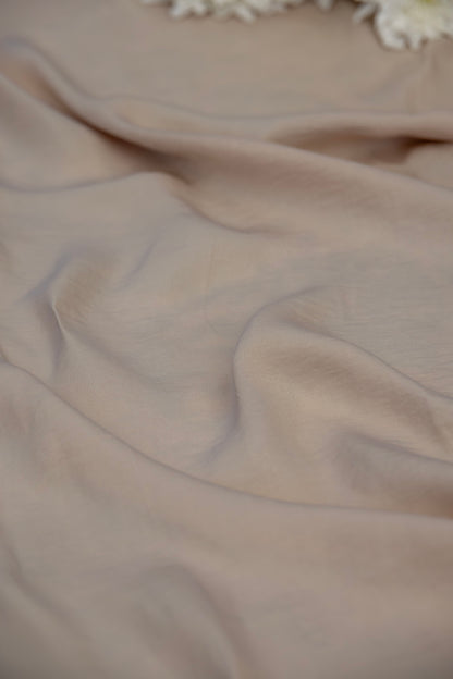 A close-up of draped light beige fabric with soft folds and a small white floral detail at the top edge.