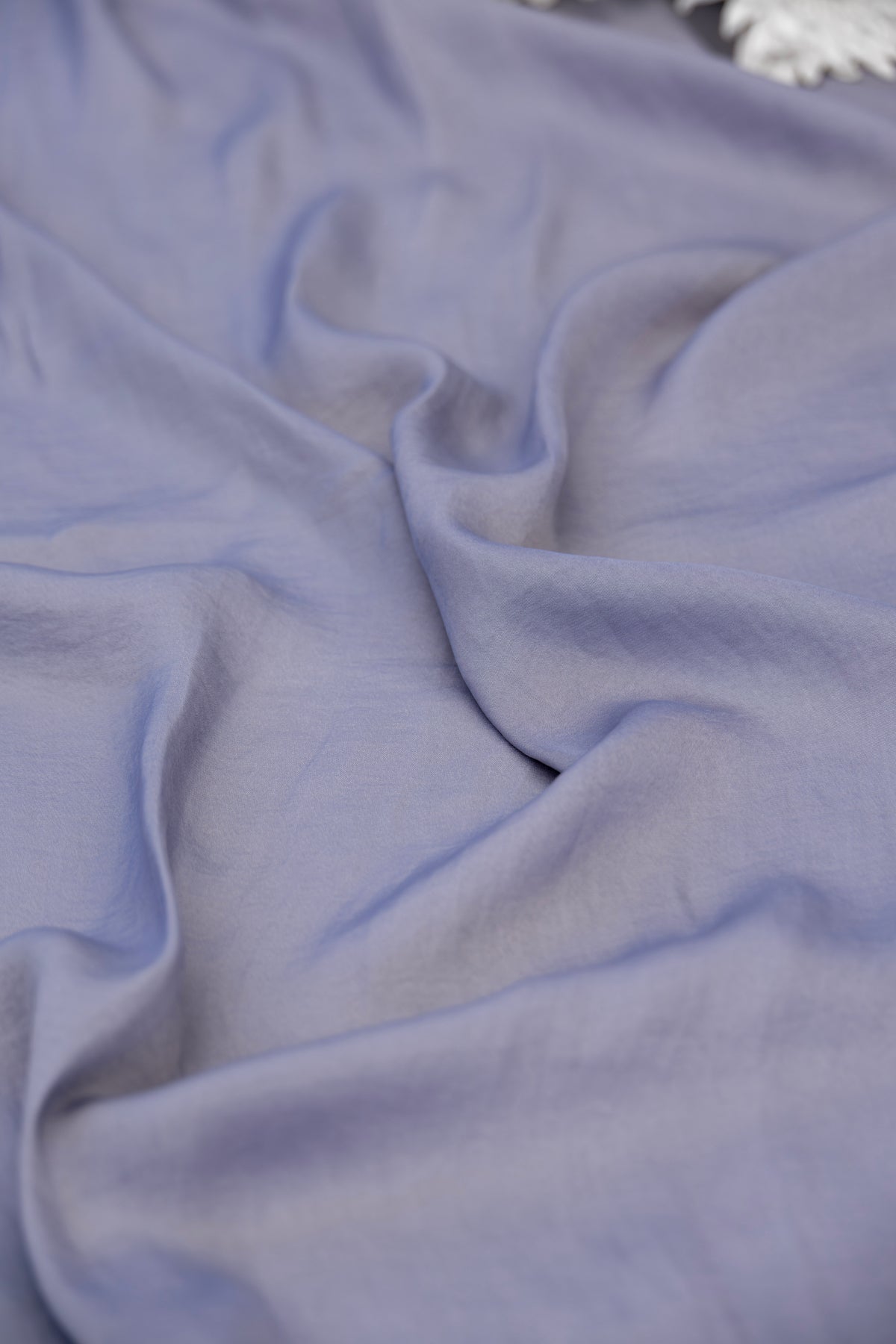 A close-up of draped light periwinkle blue fabric with soft folds and a small white decorative element visible in the top right corner.