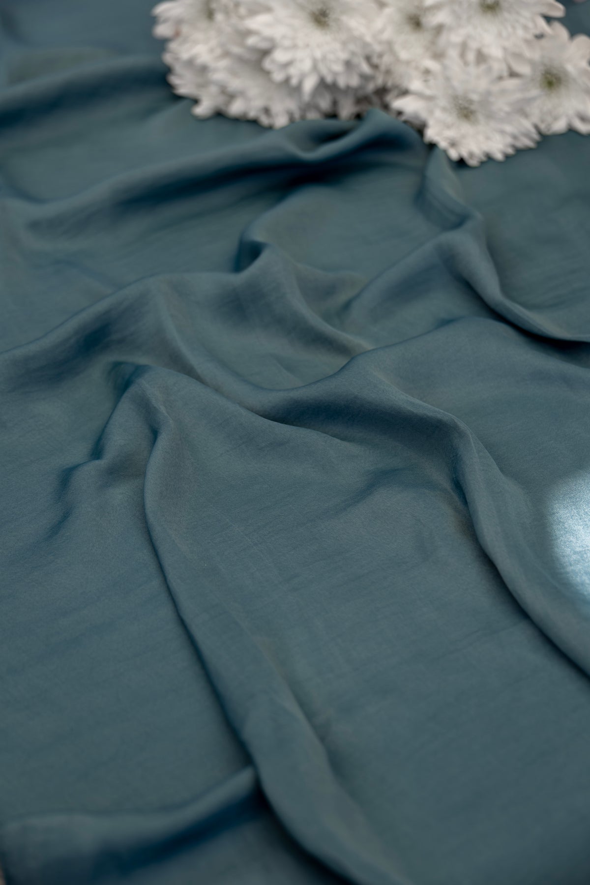 A close-up of draped slate blue fabric with soft folds, styled with white dried flowers in the upper corner.