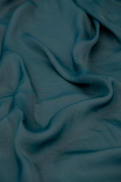 A close-up of draped teal green fabric with soft folds and a subtle woven texture.