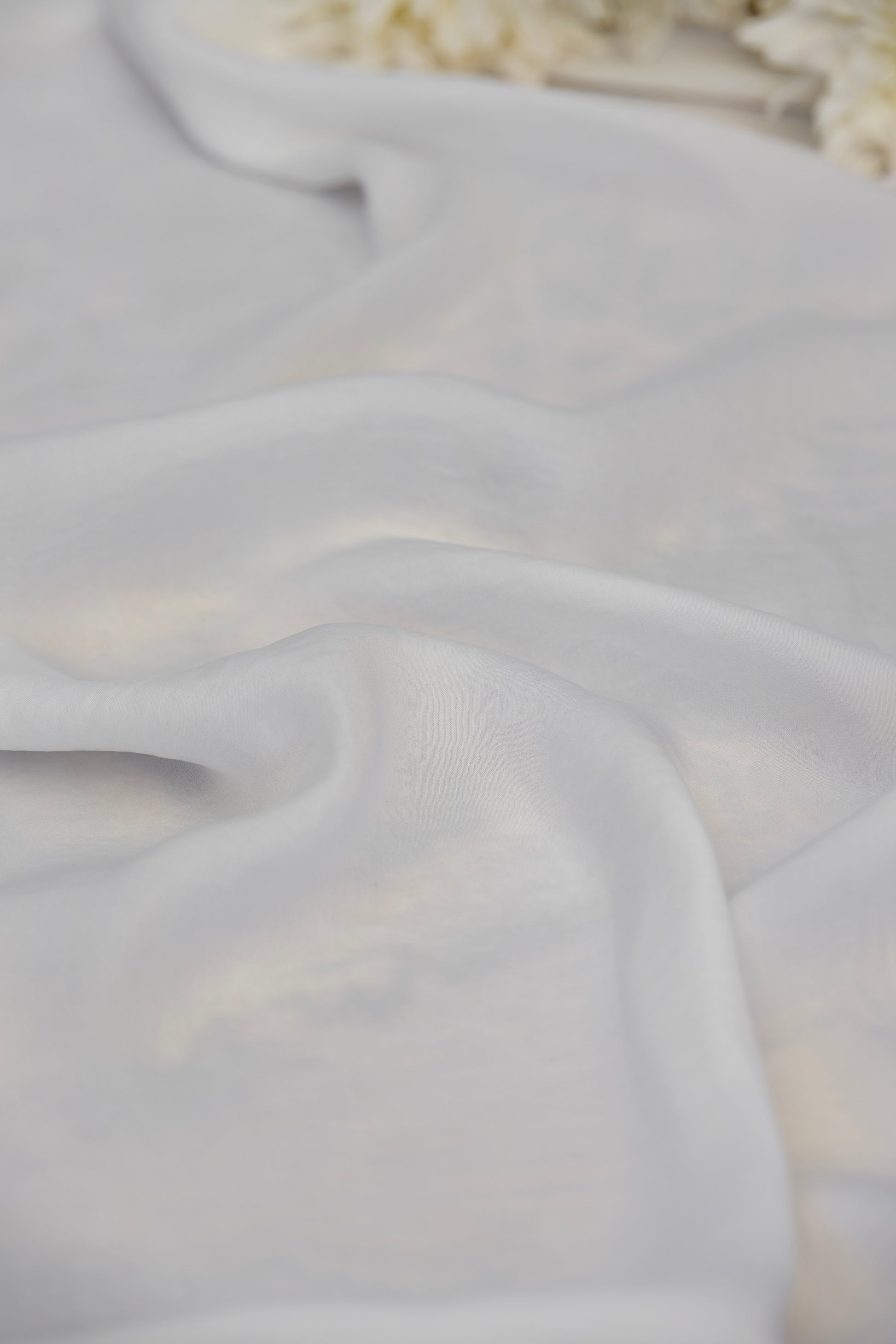 A close-up of draped white fabric with soft folds and a subtle sheen, with a blurred decorative floral element in the top right corner.