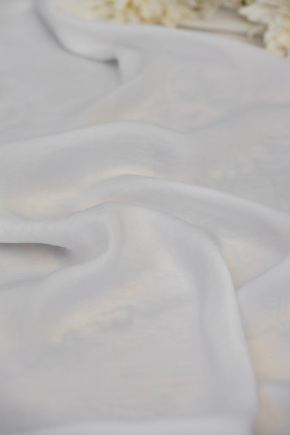 A close-up of draped white fabric with soft folds and a subtle sheen, with a blurred decorative floral element in the top right corner.