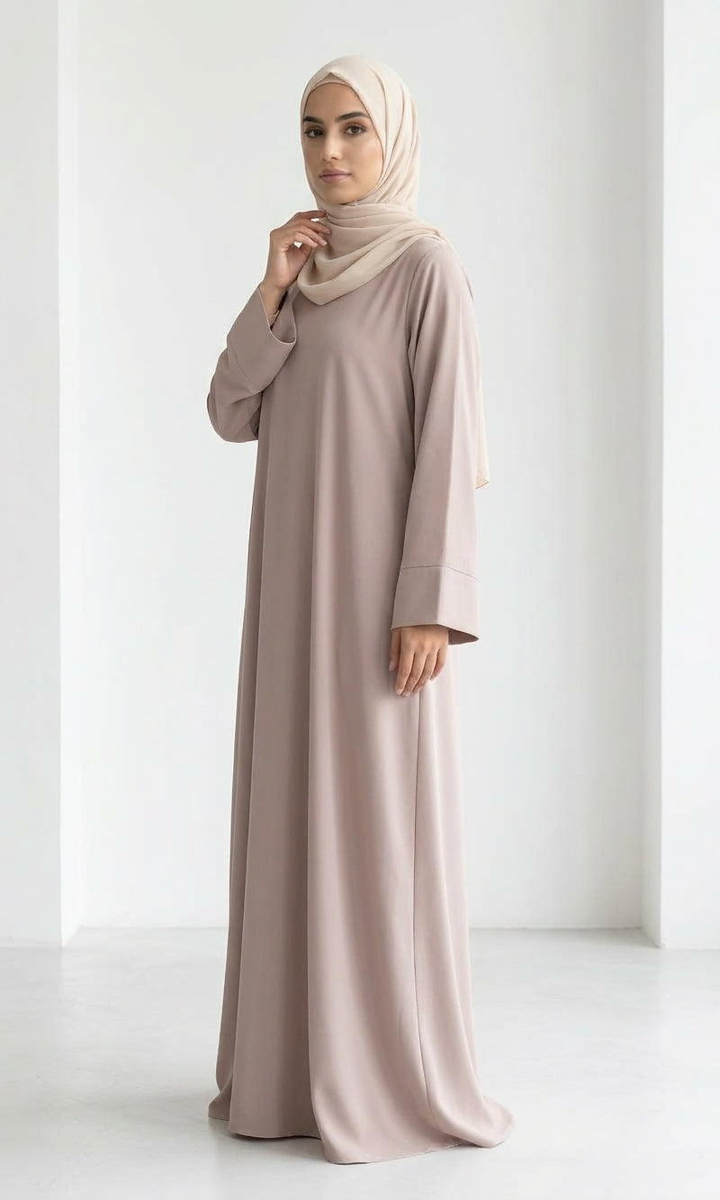 Woman wearing a long, beige colour dress and hijab standing in an indoor setting.
