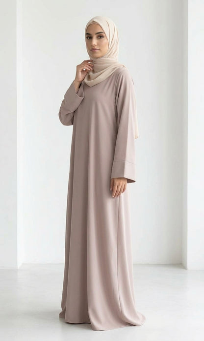 Woman wearing a long, beige colour dress and hijab standing in an indoor setting.