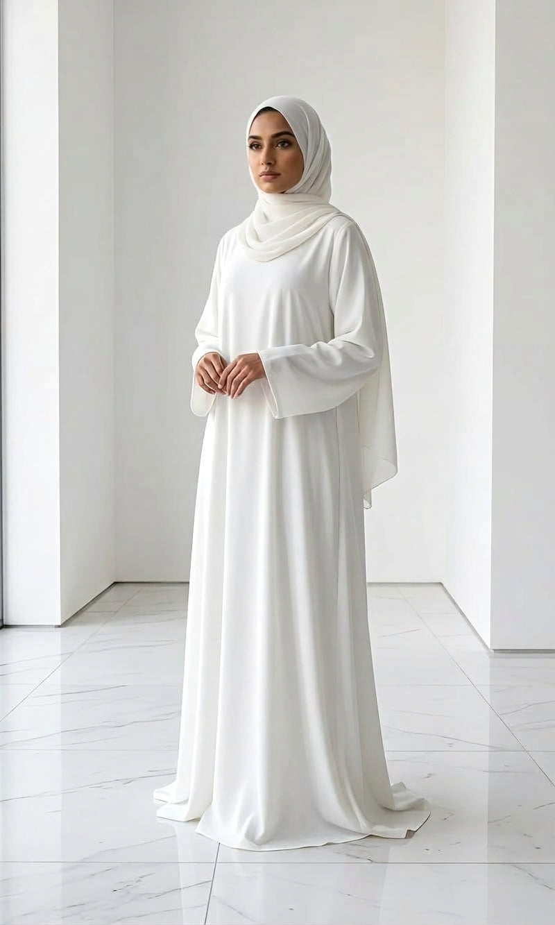 Woman wearing a long, off white colour dress and hijab standing in an indoor setting.